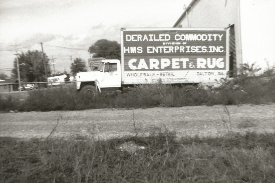 Black and white photo of a vintage delivery truck parked beside a building with large painted sign advertising “Derailed Commodity, Division of HMS Enterprises Inc. Carpet & Rug – Wholesale & Retail – Dalton, GA.