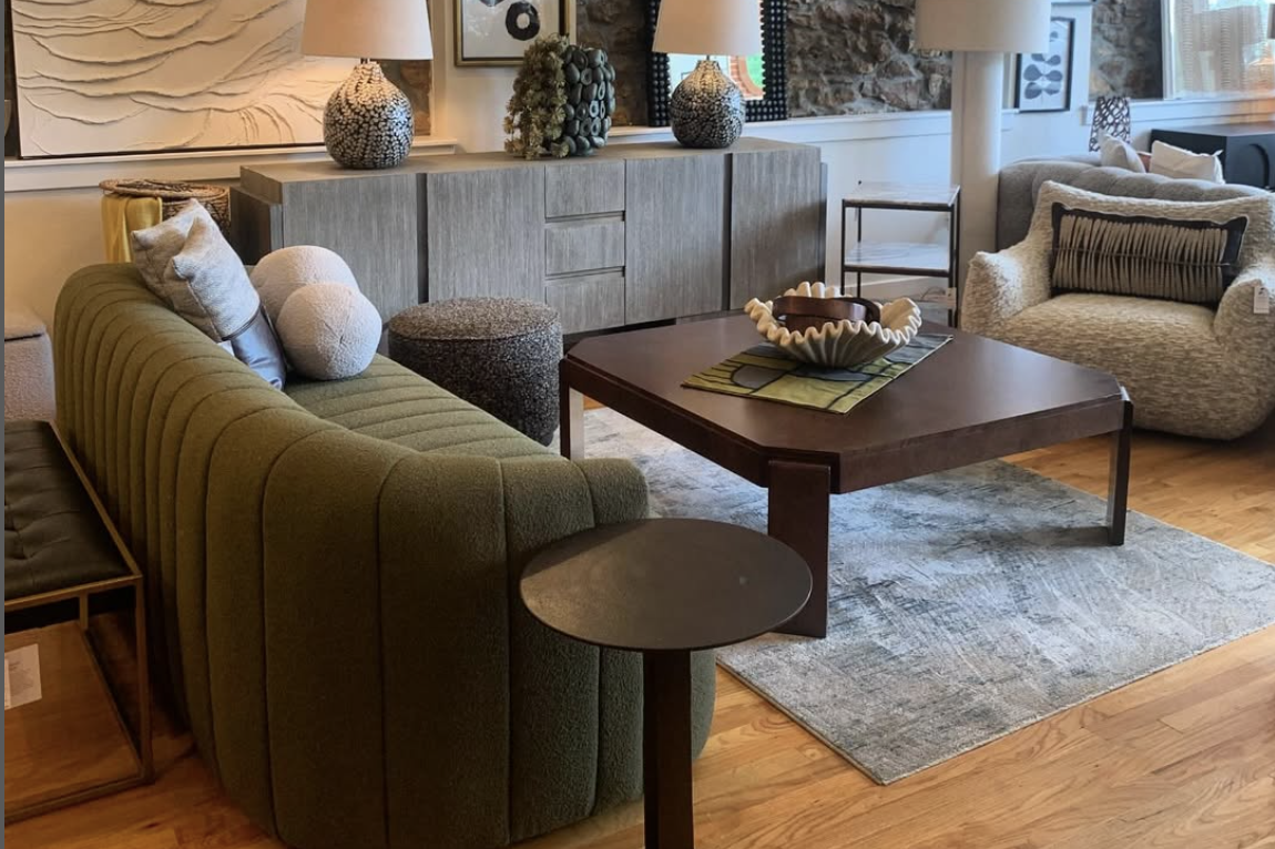Modern living room showroom with a green channel-tufted sofa, textured armchair, wood coffee table on a neutral rug, and a gray sideboard decorated with sculptural lamps and vases.