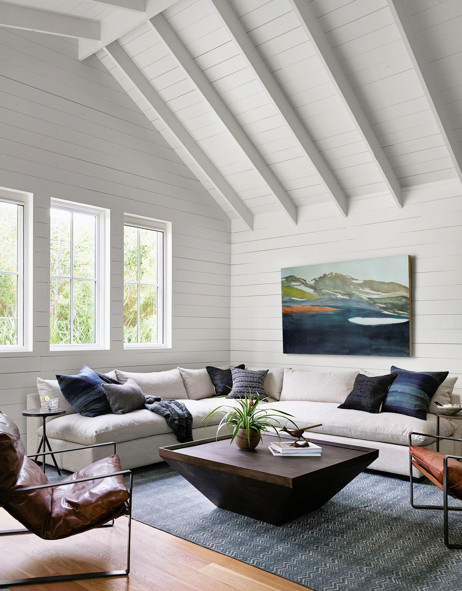 Bright modern living room with a vaulted ceiling, beige sectional sofa, dark geometric coffee table, leather accent chair, and abstract wall art, creating a cozy and contemporary space.