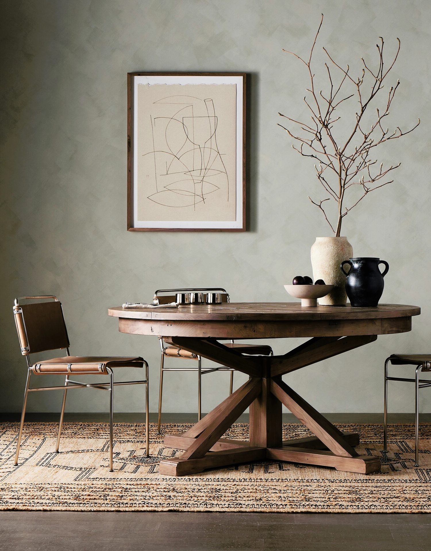 "Rustic dining room featuring a wooden round table with an X-base, paired with industrial-style metal and leather chairs, complemented by minimalist artwork and a simple vase arrangement.