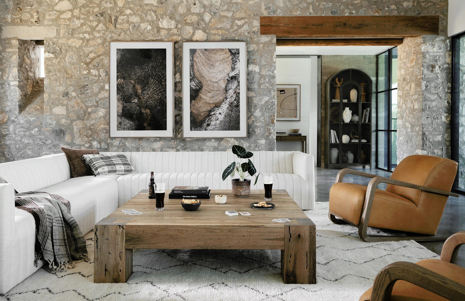 Stylish living room featuring a white sectional sofa, rustic wooden coffee table, leather armchair, and stone walls with modern artwork, creating a cozy, contemporary space.