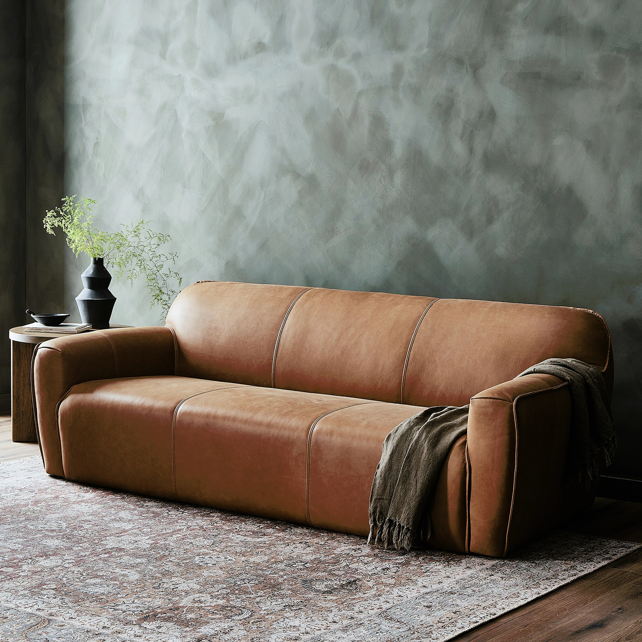 Modern camel leather sofa with plush, rounded arms and minimalist stitching, styled with a throw blanket and accent table—shop now on sale.